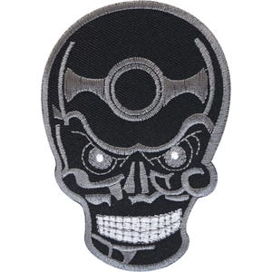 Puede incluir: Un parche bordado en negro y plata con un diseño de calavera estilizada. La calavera presenta un emblema circular en la frente, cuencas oculares angulares y una dentadura expuesta. Todo el diseño está delineado con un borde plateado.