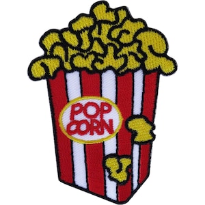 Puede incluir: Un parche bordado con forma de cubo de palomitas de maíz con granos amarillos desbordándose por la parte superior. El cubo presenta rayas verticales rojas y blancas y un óvalo blanco central con el texto rojo POP CORN bordado en el medio.