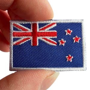 Peut inclure: Un écusson brodé rectangulaire représentant le drapeau de la Nouvelle-Zélande. Il présente un fond bleu foncé avec l'Union Jack dans le coin supérieur gauche et quatre étoiles rouges à cinq branches bordées de blanc sur la droite.
