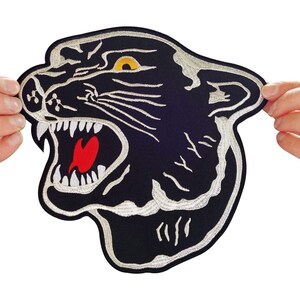 May include: An embroidered patch featuring the head of a black panther in profile with its mouth open, revealing white teeth and a red tongue. The design includes silver-grey stitching for the facial details and a bright yellow eye on a black background.