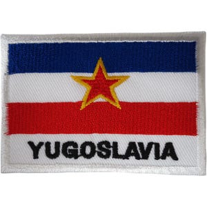 Puede incluir: Un parche bordado rectangular que muestra la bandera de Yugoslavia con franjas horizontales azul, blanca y roja. Una estrella roja con borde amarillo se sitúa en el centro. La palabra YUGOSLAVIA está bordada en letras mayúsculas negras.