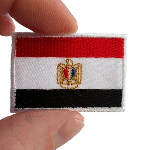 May include: A rectangular embroidered patch featuring the national flag of Egypt. The design consists of three horizontal bands in red, white, and black, with a gold-coloured eagle emblem centred on the white stripe, featuring red and blue details.