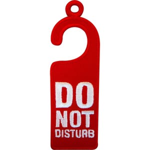 Puede incluir: Un colgador de puerta de color rojo brillante con un gancho en la parte superior para colocarlo en el pomo de una puerta. El texto DO NOT DISTURB está bordado en letras mayúsculas blancas y negritas en el centro del cuerpo rectangular.