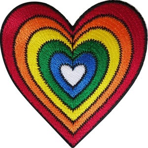 Peut inclure: Un écusson brodé en forme de cœur présentant des couches concentriques de fil dans une séquence de couleurs arc-en-ciel. Le motif commence par un centre blanc, suivi d'anneaux bleu, vert, jaune, orange et rouge, bordés d'un contour noir.