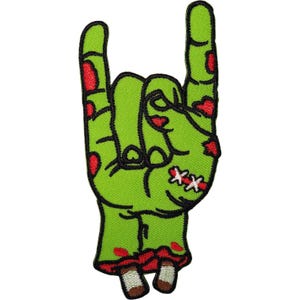 May include: An embroidered patch shaped like a severed green zombie hand making a rock and roll sign. The hand features red blood-like patches, black outlines, and white stitches on the palm, with two exposed bone ends at the wrist base.