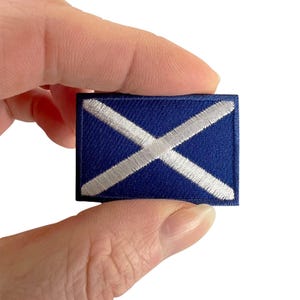 May include: A rectangular embroidered patch featuring a dark blue background with a white diagonal cross, also known as a saltire, stitched across the centre. The patch has a finished border and is held between fingers against a white background.
