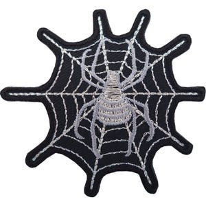 Könnte beinhalten: Ein schwarzer bestickter Aufnäher mit einer silbernen Spinne in der Mitte eines silbernen Spinnennetz-Designs. Der Aufnäher hat einen gezackten, mehrzackigen Rand und ist mit silbernem Garn auf einem schwarzen Stoffhintergrund bestickt.