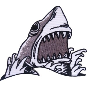 Puede incluir: Un parche bordado que muestra una cabeza de tiburón emergiendo de olas oceánicas blancas estilizadas. El tiburón está representado en tonos grises y blancos con la boca muy abierta, mostrando dientes afilados y un interior oscuro.