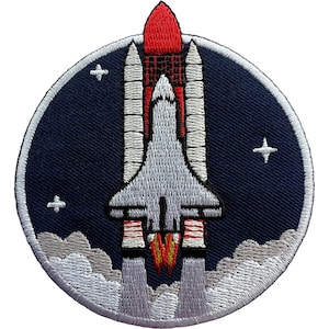 May include: A circular embroidered patch featuring a space shuttle launching into a dark blue sky. The shuttle is white with a red fuel tank and white boosters. White clouds of smoke billow at the base, and small white stars are scattered around.