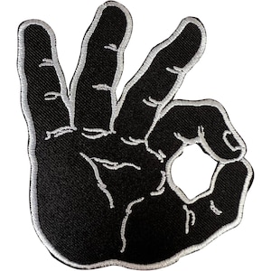 Può includere: Una toppa ricamata che raffigura la sagoma di una mano nera che fa il gesto dell'ok. Il design è delineato con filo bianco, con linee bianche cucite che dettagliano le nocche, le dita e il palmo, creando un emblema grafico ad alto contrasto.