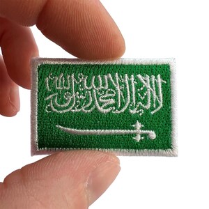 May include: A rectangular embroidered patch featuring the flag of Saudi Arabia. The design consists of a vibrant green background with white thread detailing the Arabic script of the Shahada and a white sword positioned horizontally below it.