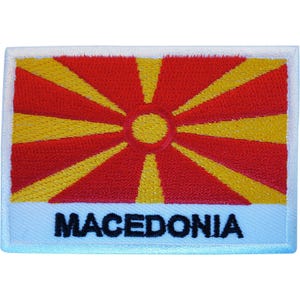 May include: A rectangular embroidered patch featuring the flag of North Macedonia with a red background and a yellow sun with eight radiating rays. The word MACEDONIA is stitched in bold black capital letters on a white band at the bottom.