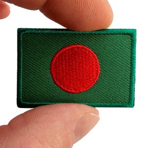 May include: A rectangular embroidered patch featuring the national flag of Bangladesh. The design consists of a solid green background with a centred, vibrant red circular emblem. The edges of the patch are finished with a thick green stitched border.