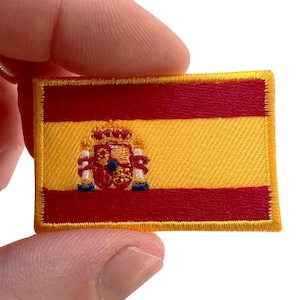 May include: A rectangular embroidered patch featuring the flag of Spain with two horizontal red stripes and a central yellow stripe. The national coat of arms is stitched in the centre of the yellow stripe using red, yellow, white, and blue thread.