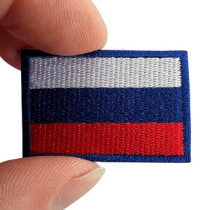 May include: A rectangular embroidered patch featuring the horizontal tricolour flag of Russia. The design consists of three equal horizontal bands of white on top, blue in the middle, and red on the bottom, all enclosed by a dark blue stitched border.