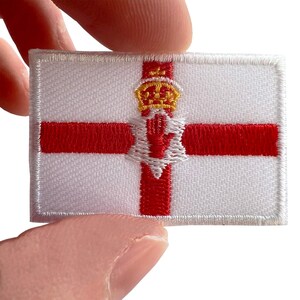 May include: A rectangular embroidered patch featuring the flag of Northern Ireland. The design consists of a white background with a red cross, a gold crown positioned above a red six-pointed star, and a red hand centred within the star.