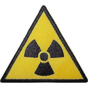 Op de afbeelding: Een driehoekige geborduurde patch met een felgele achtergrond en een zwarte rand. In het midden is een zwart stralingsgevaarsymbool afgebeeld, bestaande uit een centrale cirkel omringd door drie gestileerde waaiervormige bladen.