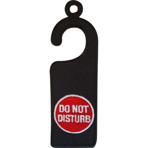 Könnte beinhalten: Ein schwarzer, bestickter Türhänger mit einem Haken an der Oberseite. Der untere Teil zeigt einen kreisförmigen roten Aufnäher mit weißem Rand, der den weißen, gestickten Text DO NOT DISTURB in einer fetten serifenlosen Schrift enthält.