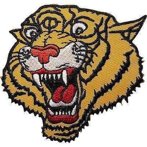 May include: An embroidered patch featuring the head of a tiger with yellow fur, black stripes, a red nose, and a wide-open mouth showing white teeth and a red tongue. The design is outlined in black thread with a jagged edge and white accents.