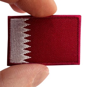 May include: A rectangular embroidered patch featuring the flag of Qatar. The design consists of a maroon field on the right side and a white vertical band on the left side, separated by a vertical line of nine white triangular serrated points.