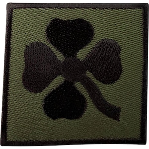 Könnte beinhalten: Ein quadratischer, bestickter Aufnäher mit olivgrünem Hintergrund und einem schwarzen vierblättrigen Kleeblatt-Motiv in der Mitte. Der Aufnäher ist mit einem dicken, schwarz gestickten Rand versehen, der die gesamte quadratische Kante einfasst.