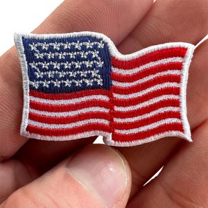 Puede incluir: Un parche bordado rectangular que presenta la bandera estadounidense con un diseño ondulado. El parche muestra un campo azul con estrellas blancas bordadas a la izquierda y rayas horizontales rojas y blancas alternas a la derecha, con un borde blanco.