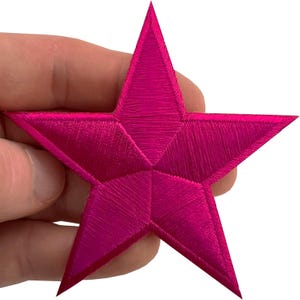Pode incluir: Um emblema em forma de estrela de cinco pontas com bordado rosa choque vibrante e um brilho distinto. O design inclui linhas de fio visíveis que irradiam do centro para cada ponta, criando uma aparência geométrica texturizada.