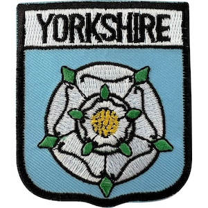 Puede incluir: Un parche bordado en forma de escudo con fondo azul claro y borde negro. En la parte superior, la palabra YORKSHIRE está bordada en negro sobre un campo rectangular blanco. Debajo, una rosa blanca con sépalos verdes y centro amarillo.