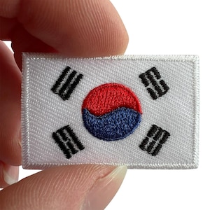 Puede incluir: Un parche rectangular bordado con el diseño de la bandera de Corea del Sur. El parche tiene un fondo blanco con un símbolo taegeuk rojo y azul en el centro y cuatro trigramas negros en cada esquina, todo acabado con un borde blanco.
