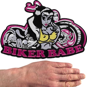 May include: An embroidered patch featuring a stylized figure with black hair wearing a yellow top, positioned in front of a motorcycle. The design includes pink outlines and the text BIKER BABE in pink lettering at the bottom. The patch is shown above a hand.