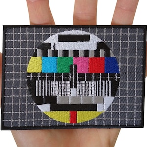 May include: A rectangular embroidered patch featuring a classic television test pattern. The design includes a circular center with vibrant yellow, cyan, green, magenta, red, and blue color bars set against a dark gray grid background.
