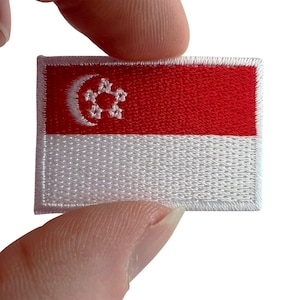 Può includere: Una toppa ricamata rettangolare raffigurante la bandiera di Singapore. Il design è composto da una fascia orizzontale rossa nella parte superiore e una bianca in quella inferiore. La sezione rossa include una mezzaluna e cinque stelle bianche.