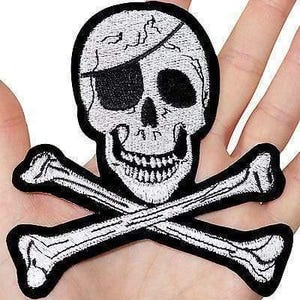May include: An embroidered patch featuring a white skull with a black eye patch and a black nose, positioned above two crossed white bones. The entire design is outlined in a thick black border, creating a high-contrast pirate-themed emblem.
