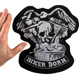 Op de afbeelding: Een zwarte geborduurde patch met een gedetailleerde grijze en witte afbeelding van een V-twin motorblok met een centraal schedelontwerp. Onder de motor bevindt zich een gebogen banner met de witte geborduurde tekst BIKER BORN in hoofdletters.
