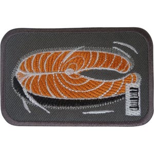 Op de afbeelding: Een rechthoekige geborduurde patch met een donkergrijze achtergrond met een gestileerde oranje zalmmoot met witte details. Een klein wit rechthoekig icoon met zwarte verticale lijnen die op pianotoetsen lijken, bevindt zich rechtsonder.