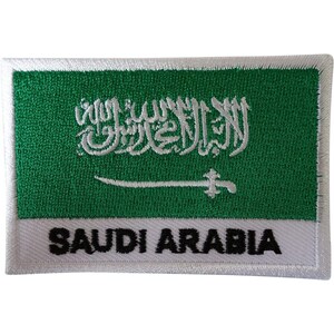 May include: A rectangular embroidered patch featuring the flag of Saudi Arabia with a green background, white Arabic calligraphy, and a white sword. Below the flag, the text SAUDI ARABIA is embroidered in bold black capital letters on a white border.
