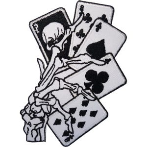May include: An embroidered patch featuring a skeletal hand holding a fan of playing cards. The design is rendered in black and white thread, showcasing a skull on the top card, followed by clubs, a spade, and diamonds on the other cards.