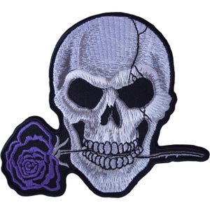 May include: An embroidered patch featuring a light gray human skull with a visible crack on the right side and deep black eye sockets. The skull holds a single purple rose with dark outlines in its teeth, all bordered by a thick black edge.