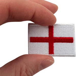 Puede incluir: Un parche bordado rectangular que presenta la bandera de Inglaterra, la cual consiste en un fondo blanco con una cruz roja centrada. El bordado tiene un patrón de hilo texturizado visible en todo el diseño con un borde cosido.