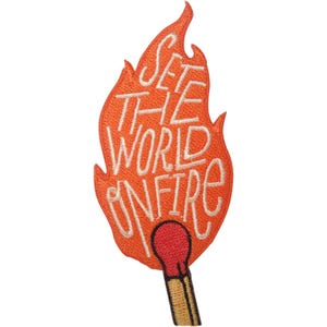 Puede incluir: Un parche bordado con forma de llama y una cerilla en la base. La llama naranja contiene el texto bordado en blanco "SET THE WORLD ON FIRE". La cerilla tiene una cabeza roja y un tallo de madera color canela con detalles de contorno marrón.