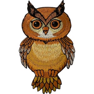 Puede incluir: Un parche bordado de un búho con hilos en tonos marrón, beige y naranja. El búho tiene ojos grandes, oscuros y redondos, un pequeño pico puntiagudo y plumas detalladas en las alas. Todo el diseño está delineado con un borde negro grueso.