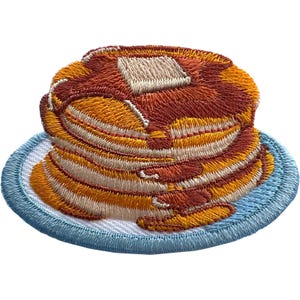 May include: An embroidered patch featuring a stack of golden-brown pancakes topped with a square pat of butter, resting on a light blue plate. The design uses shades of brown, orange, and cream thread to create a textured, detailed food illustration.