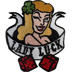 Op de afbeelding: Een geborduurde patch met een blonde vrouw met een groen klaverblad in haar haar, die knipoogt. Een witte banner in het midden toont de tekst LADY LUCK in zwarte letters. Twee rode dobbelstenen met zwarte stippen staan onderaan.