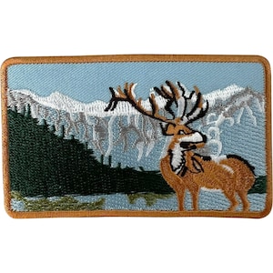 May include: A rectangular embroidered patch featuring a brown elk with large antlers standing in front of a dark green forest and snow-capped mountains against a light blue sky. The patch has a thick, solid brown stitched border.