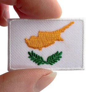 May include: A rectangular embroidered patch featuring the flag of Cyprus. The design consists of a white background with a golden-orange map of the island of Cyprus centered above two crossed green olive branches, all finished with a white border.
