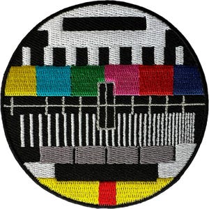 Op de afbeelding: Een ronde geborduurde patch met een klassiek televisie-testbeeld. Het ontwerp bevat een horizontale rij met gele, cyaan, groene, magenta, rode en blauwe kleurenbalken, geplaatst tegen een zwarte achtergrond met witte geometrische lijnen.