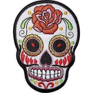 May include: An embroidered patch shaped like a skull with a white base and black border. The design features a large orange rose on the forehead, yellow flower-shaped eye sockets with black centres, red cheek markings, and a pink-outlined toothy grin.