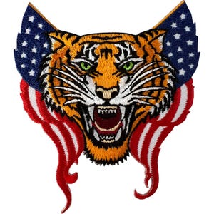 Könnte beinhalten: Ein gestickter Aufnäher mit einem knurrenden Tigerkopf, leuchtend grünen Augen, orange-schwarzem Fell und weißen Schnurrhaaren. Der Tiger ist von einem stilisierten amerikanischen Flaggendesign mit blauen Bereichen und weißen Sternen umrahmt.