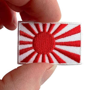 May include: A rectangular embroidered patch featuring a white background with a central red circle and sixteen red rays extending outward to the edges. The design is stitched with a dense thread texture and finished with a white merrowed border.