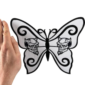 Könnte beinhalten: Ein weißer, schmetterlingsförmiger Stickaufnäher mit schwarzen Umrandungen und zwei detaillierten Totenkopfmotiven mit kleinen Hörnern auf jedem Flügel. Der Aufnäher hat ein symmetrisches Design mit einem schwarzen Mittelkörper.